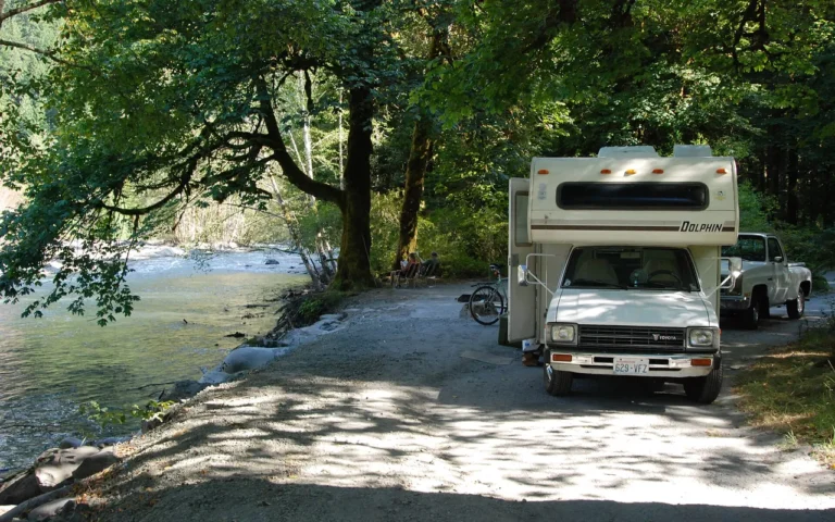 A small motorhome on a shaded campground road beside a lake